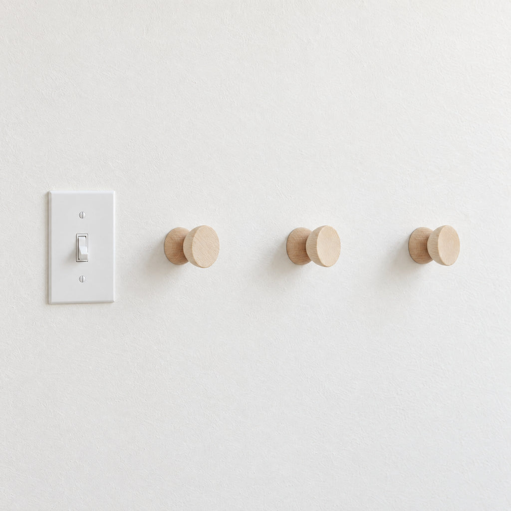 Three round beech wood hooks in a row beside a light switch on a white wall