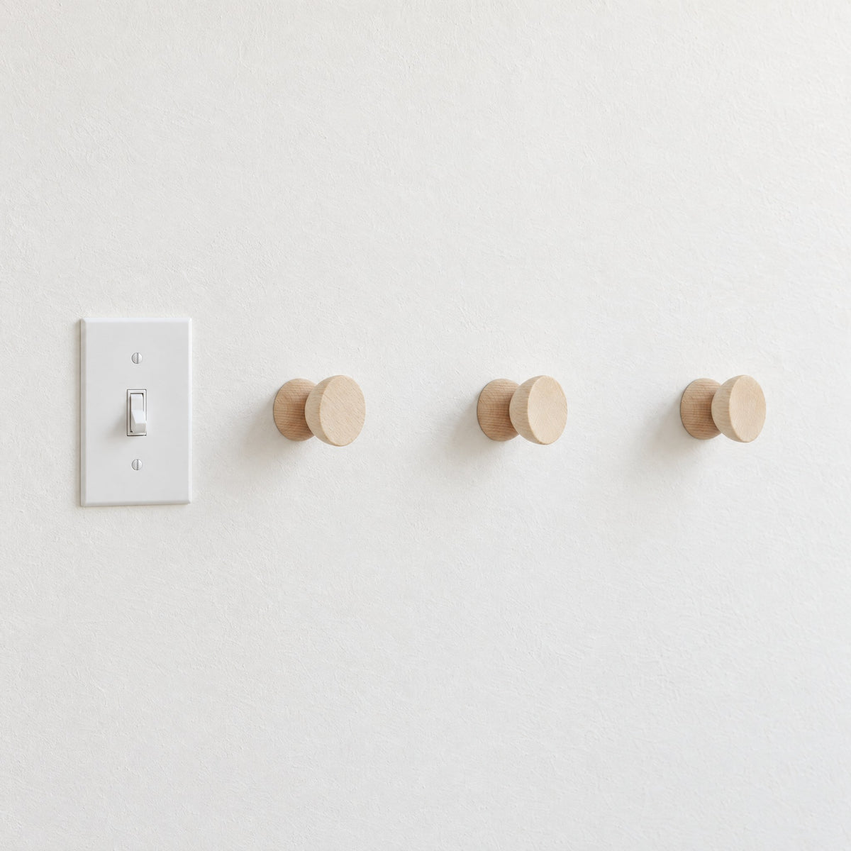 Three round beech wood hooks in a row beside a light switch on a white wall