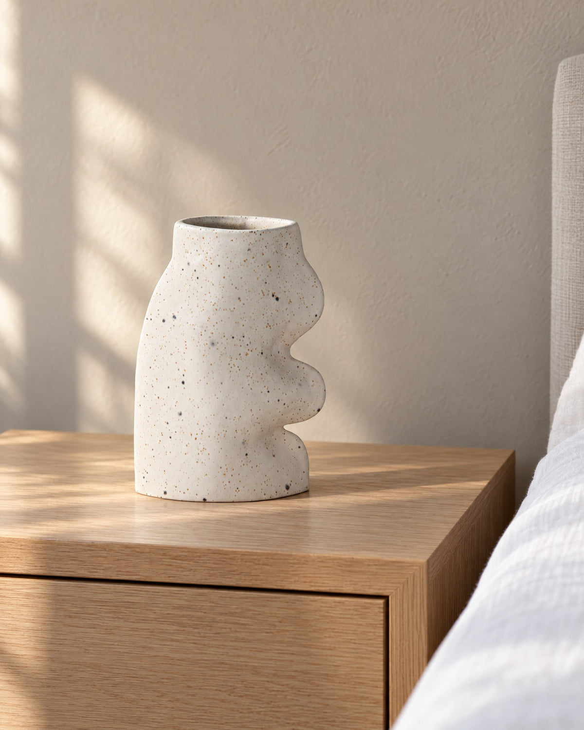 Fluxo large white speckled ceramic vase, empty, on a light oak nightstand beside a bed in morning light