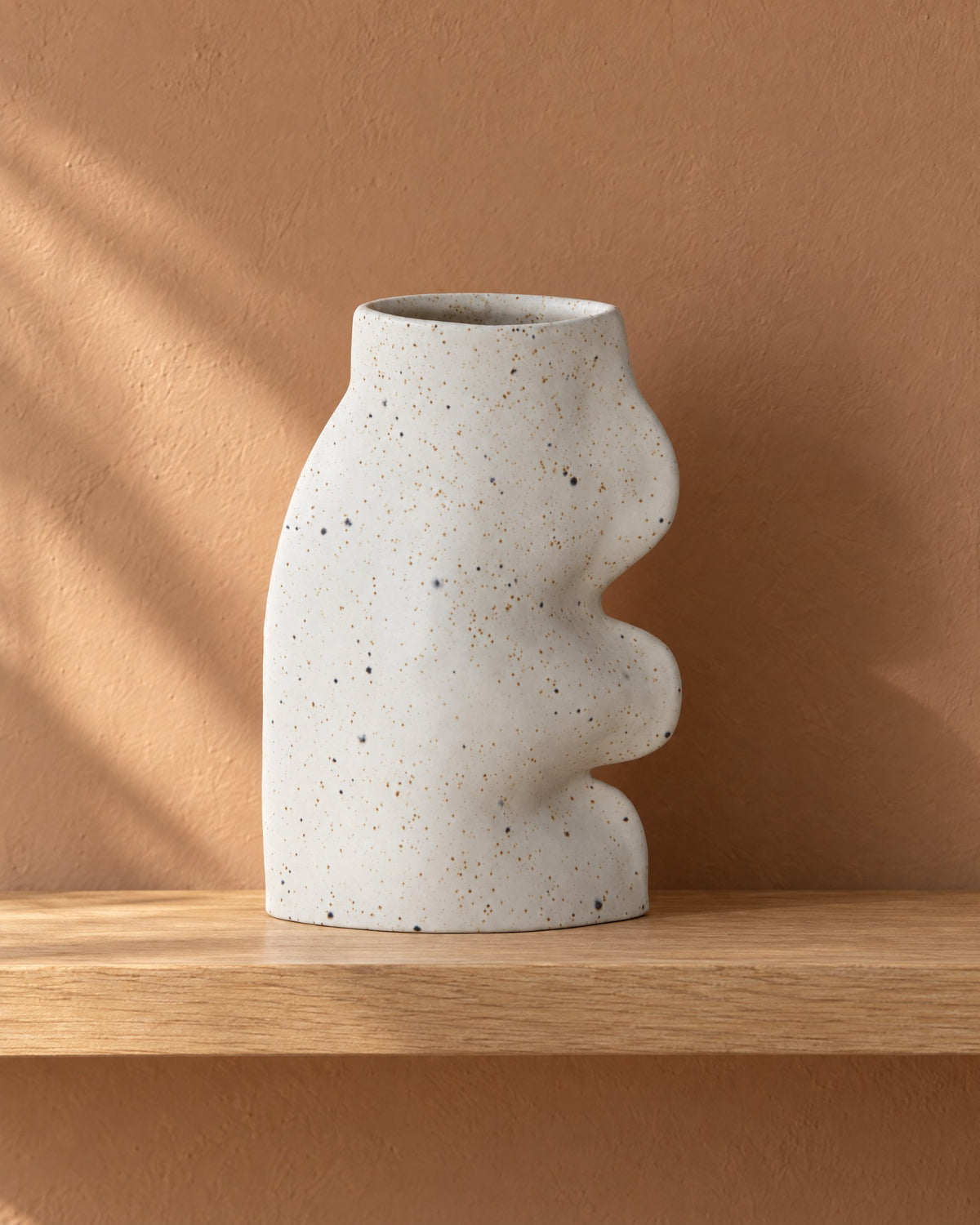 Fluxo large white speckled ceramic vase, empty, on a light oak wall shelf against a clay plaster wall