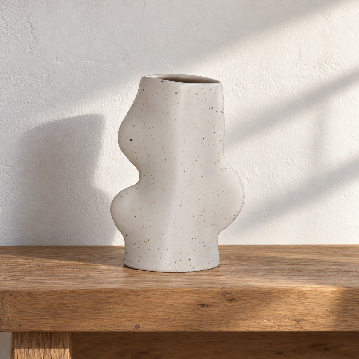 Fluxo medium white speckled ceramic vase, empty, on an oak bench against a white limewash wall in afternoon sun