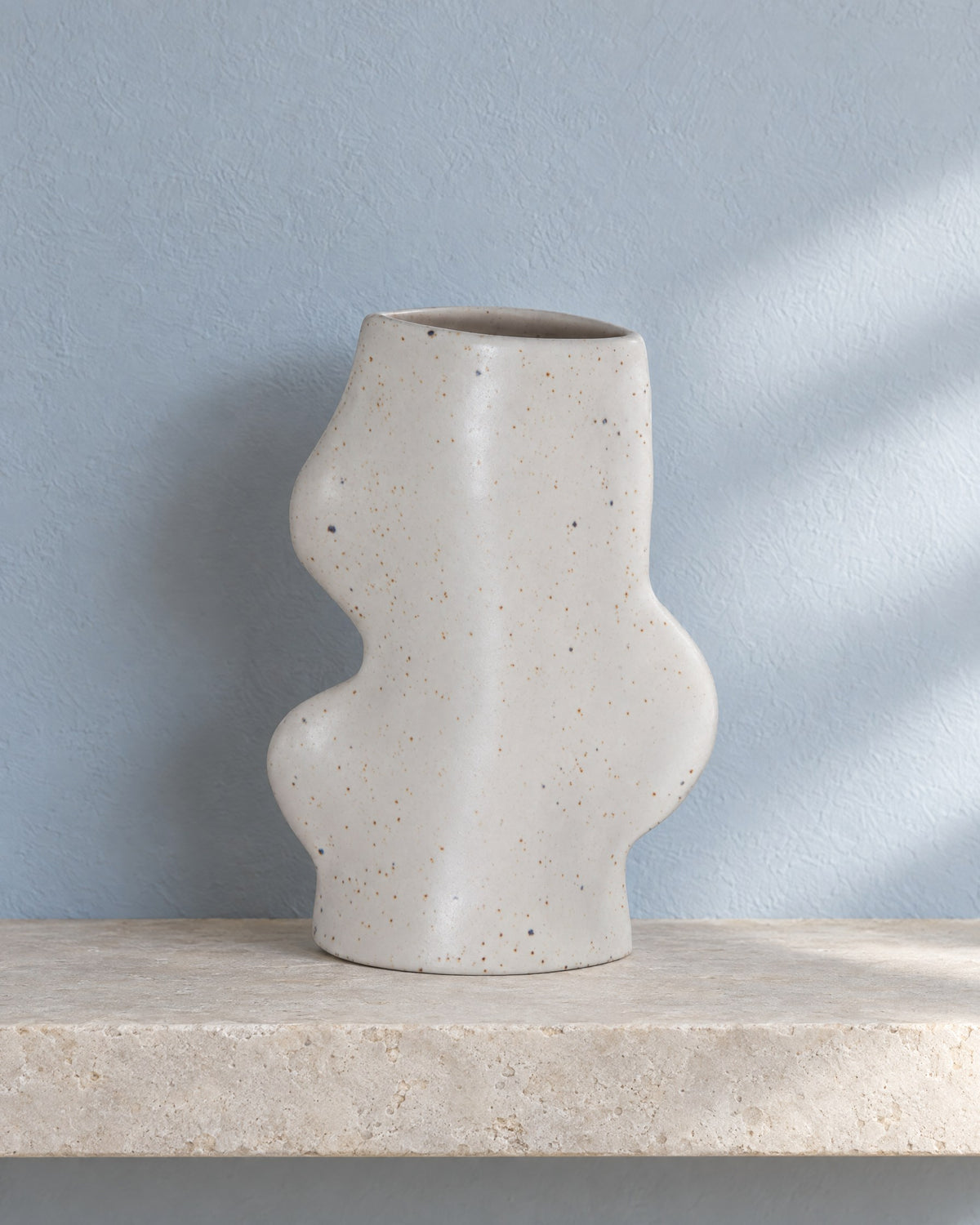Fluxo medium white speckled ceramic vase, empty, on a limestone wall shelf against a blue-grey plaster wall