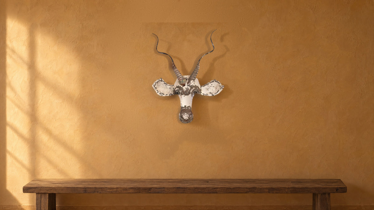 Kudu recycled metal wall mask with spiral horns on an ochre plaster wall above a long dark oak bench, in late-afternoon light