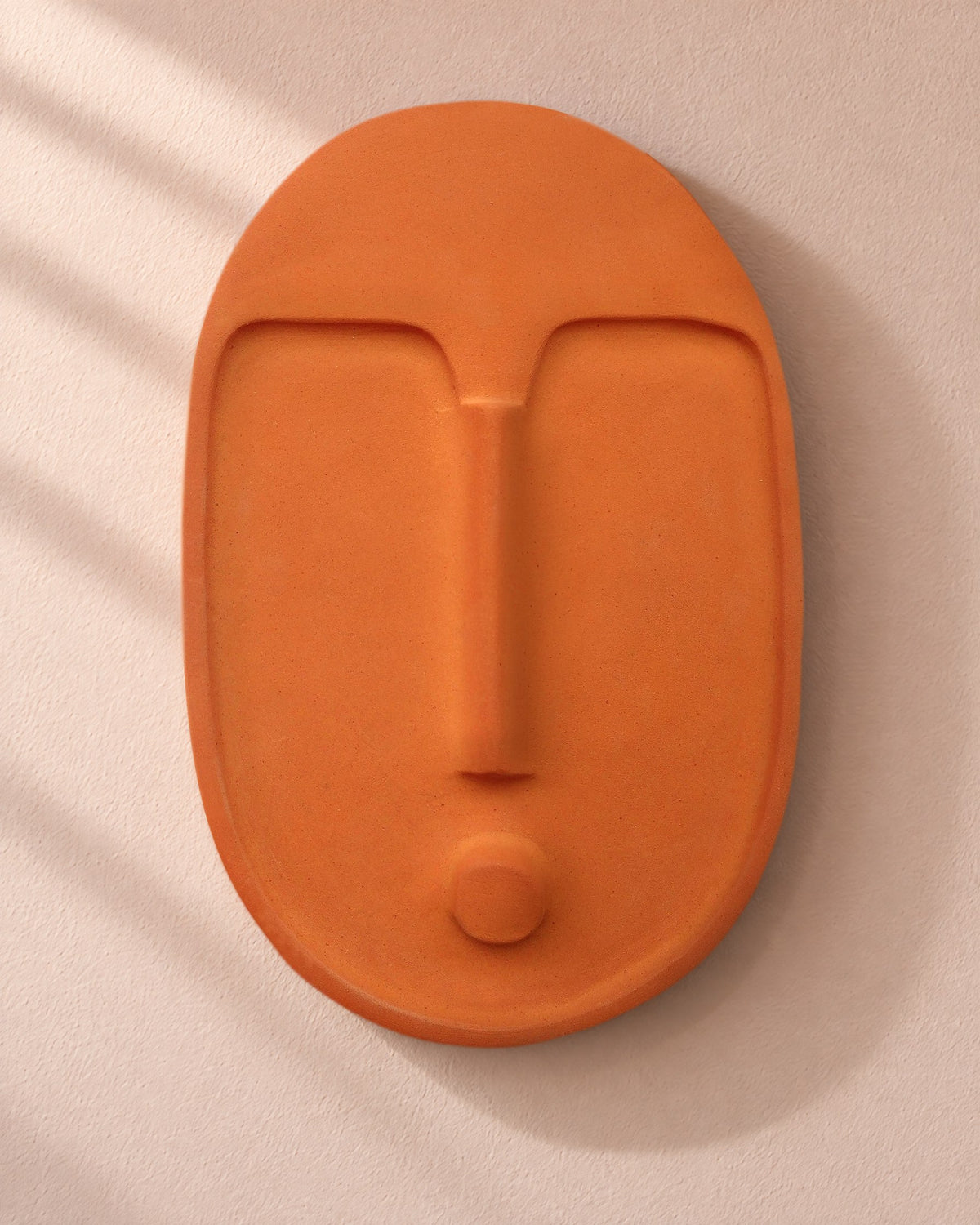 The terracotta ceramic wall mask, front view, on a blush plaster wall in raking window light