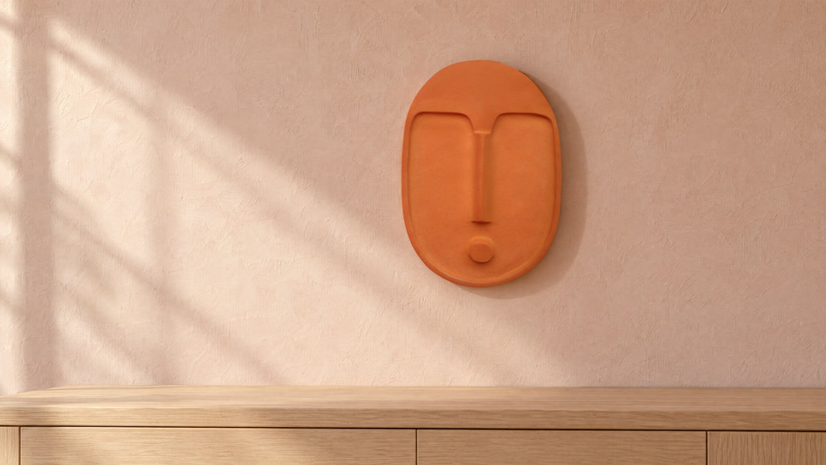 Terracotta ceramic wall mask on a blush-pink plaster wall above a pale oak sideboard, in late-morning window light