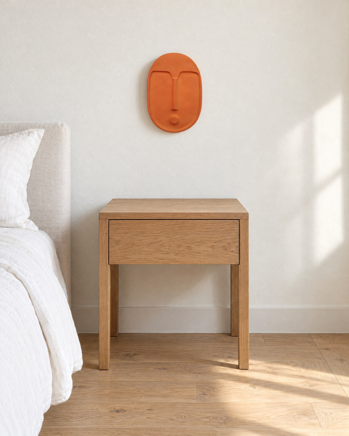 Terracotta ceramic wall mask hung above a small oak nightstand in a white bedroom