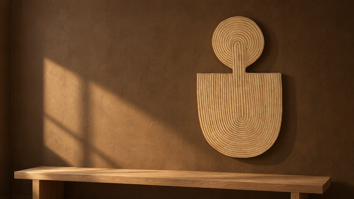Abstract Wall Mask 1 hung on a dark umber plaster wall in a beam of late sun, above a long low oak bench