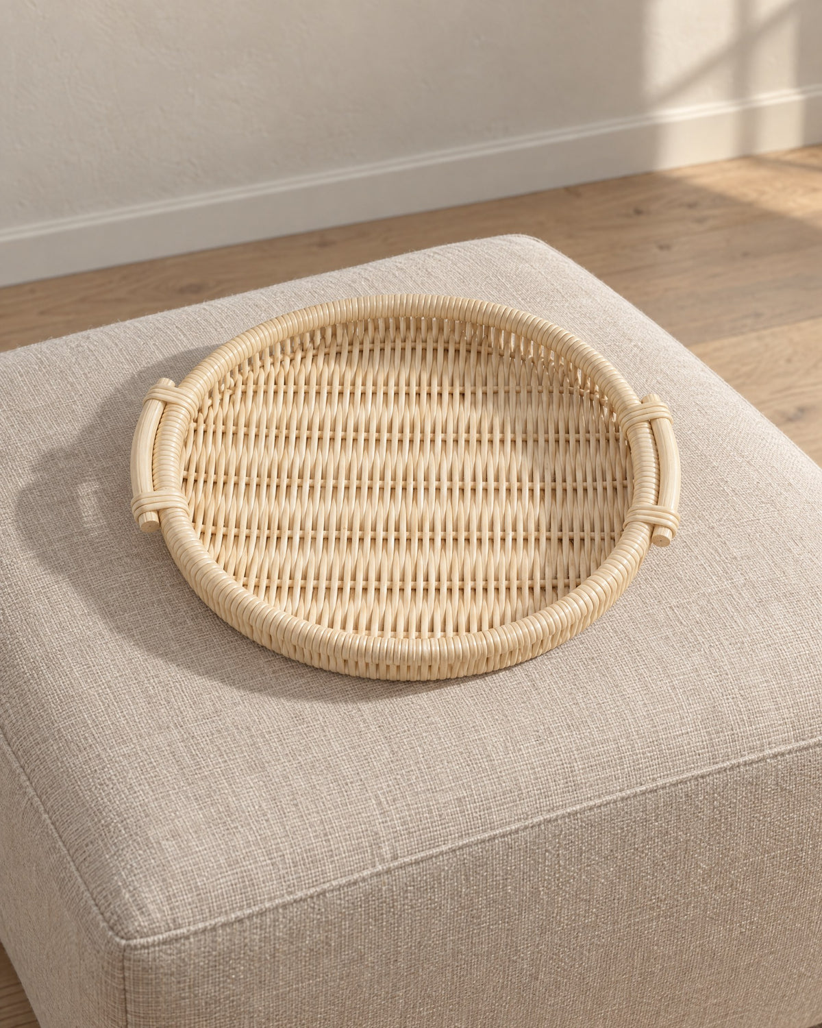 Round natural woven rattan tray, empty, on an oatmeal linen ottoman in afternoon light