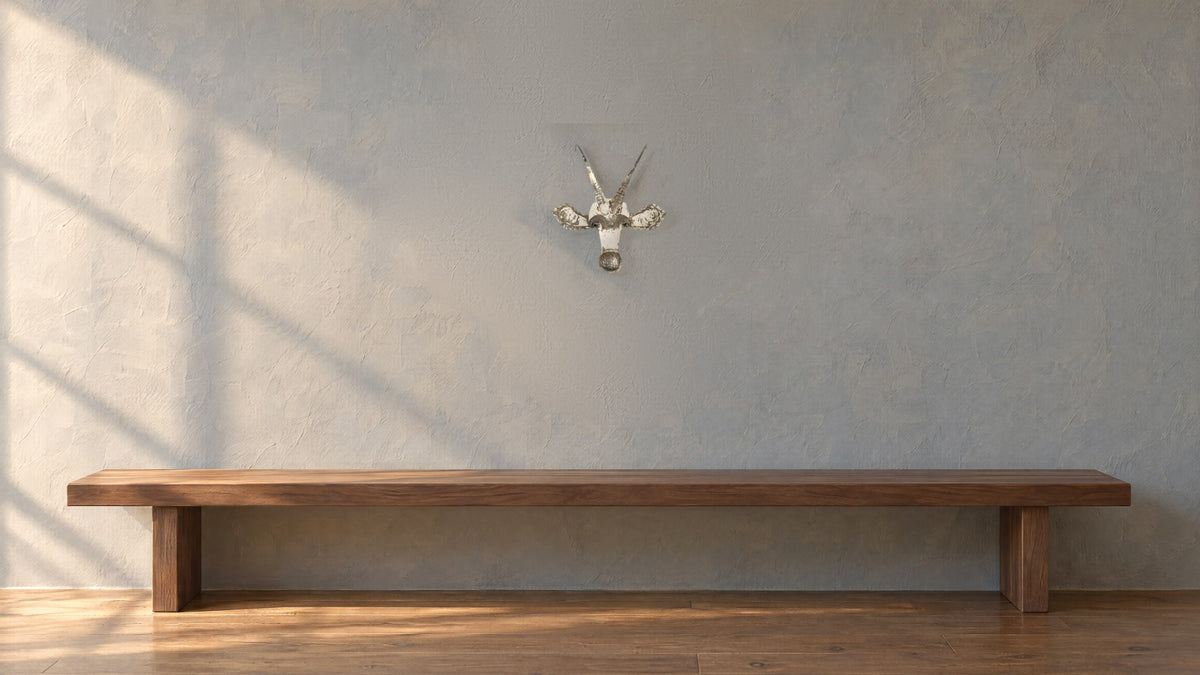 Sable recycled metal wall mask with two long horns on a grey-blue plaster wall above a long low walnut bench, in late-afternoon light