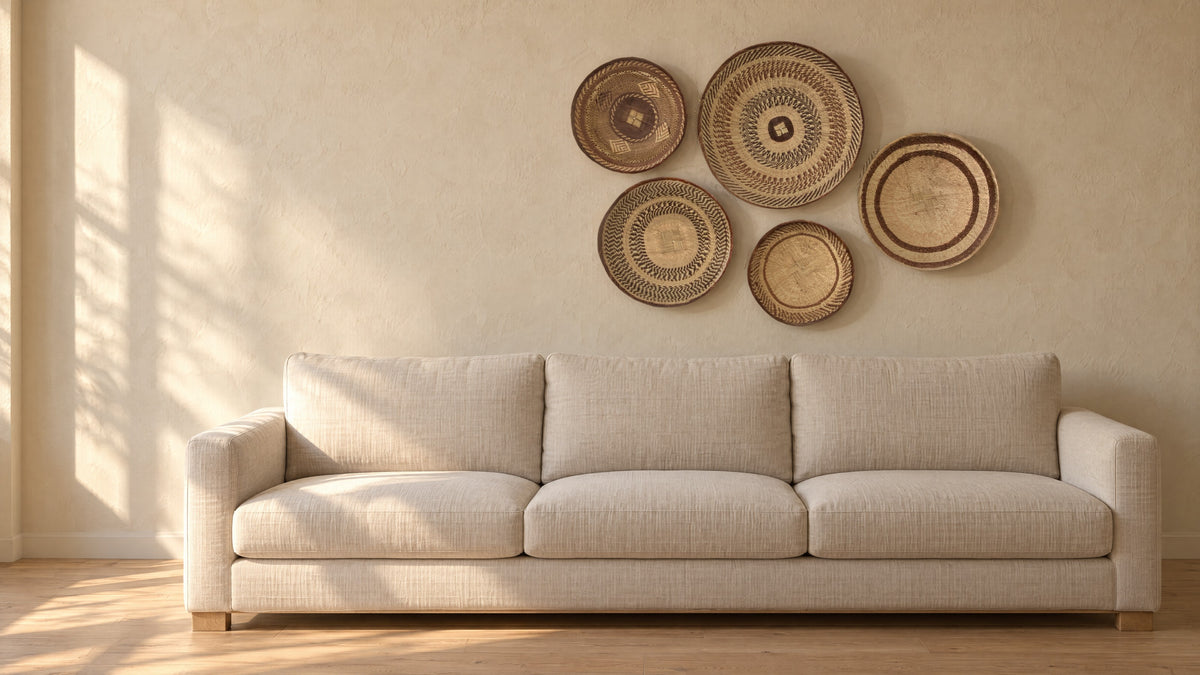 Five woven Tonga baskets hung as a group on a warm sand plaster wall above a plain linen sofa, in late-afternoon sun
